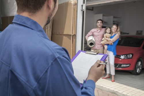 The image shows a man in a blue uniform standing outside a residential garage, holding a clipboard and pen, likely organizing a house move for Streatham Man with Van. Inside the garage, there are several cardboard boxes stacked to the left, some with visible tape and labels, indicating they are packed for relocation. To the right, a red car is parked inside the garage, partially visible with visible headlights and front grille, suggesting it is parked for a move or collection. In the background, a smiling young family — man, woman, and a small girl — stand on a driveway near the garage entrance, with the man holding a rolled-up carpet or mat, and the woman embracing the girl. The exterior is part of a suburban residential street, with a concrete driveway and the garage door slightly open, revealing a tidy indoor space with shelves or storage units faintly visible. Natural daylight illuminates the scene, creating a professional, organized atmosphere typical of a home removals service, subtly conveying local expertise in Streatham or nearby London postcode areas.