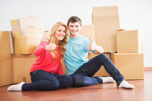 A young couple sitting on a wooden floor in a well-lit room, surrounded by stacked cardboard boxes of various sizes, indicating a house relocation. The woman has long blonde hair and is wearing a red top and dark jeans, while the man has short dark hair and is dressed in a light blue top and dark jeans. They are smiling, giving a thumbs-up, and appear to be happy with their move. The background shows a plain light-colored wall, and the boxes are arranged casually with some on top of each other, suggesting an active packing or unpacking process typical of local house removals. The scene conveys a positive, organized atmosphere consistent with a professional house removals service such as Streatham Man with Van, operating in the Streatham area, possibly within the SW postcode. The overall setting emphasizes the domestic nature of the move and the efficiency of local removals in the area.
