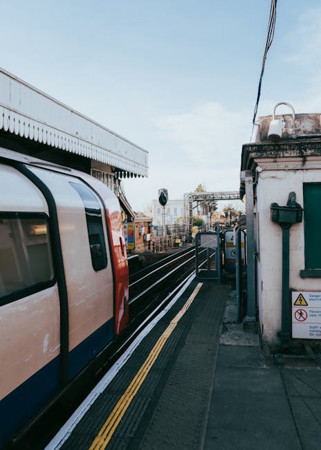 A suburban train arriving at a station platform with a white and red exterior, positioned on railway tracks next to a small building with a green window and a black mailbox mounted on the wall. The pl