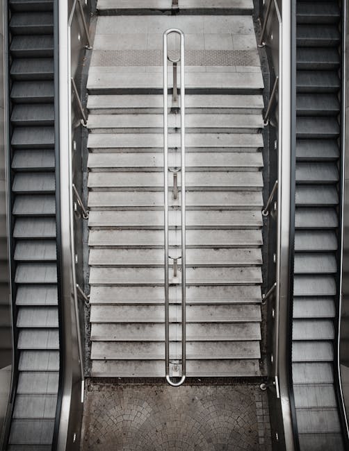 A top-down view of a set of metallic stairs with horizontal slip-resistant treads, leading down to a landing paved with small, light-colored tiles. The stairs are flanked by two escalators with black